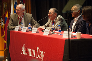 Photo of three men at a panel with the table banner displaying "Alumni Day"