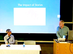 Man sits at a table in front of a presentation screen that reads The Power of Storytelling, while Neal Baer stands at a podium.