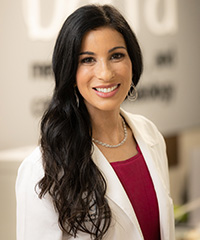 Woman with long, dark hair wearing a white coat smiling at the camera.