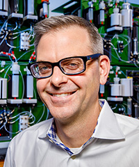 Man wearing glasses and a collared shirt in front of a molecule making machine.
