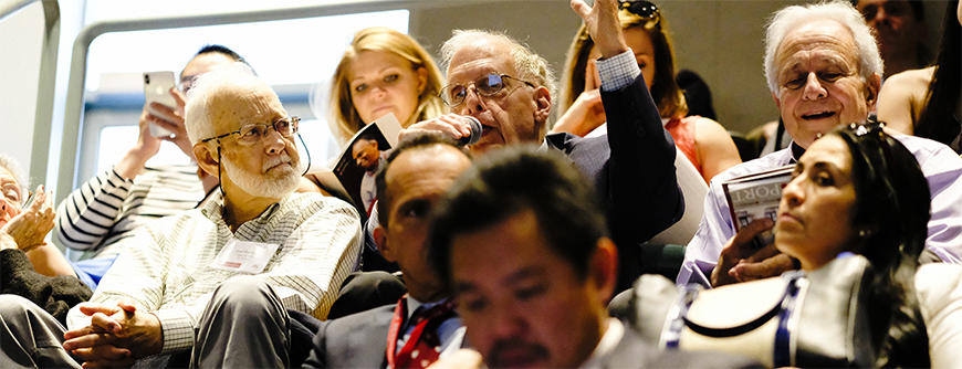 Photo of a crowd of people sitting in an auditorium listening to a talk. One man is holding a microphone and asking a question.