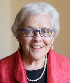 Eleanor Gossard Shore smiling wearing blue glasses, a red coat, and pearls. 